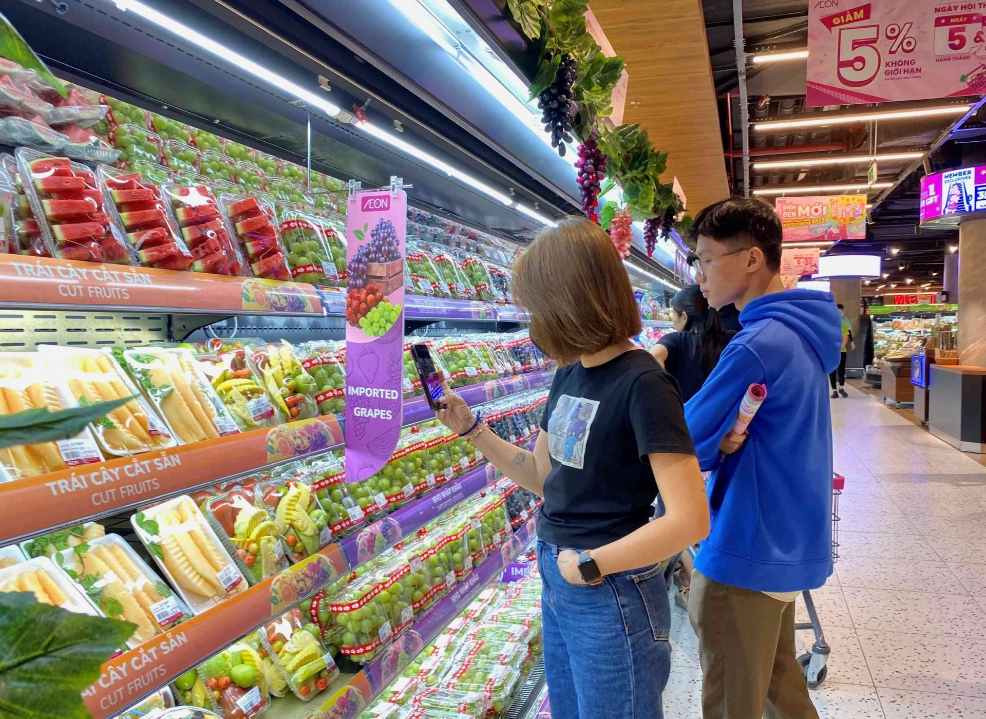 Shoppers browsing a refrigerated produce section with packaged fruits and a prominent “Imported Grapes” promotional banner at AEON supermarket.